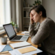 Freelancer met hoofd in hand aan minimalistische thuiswerkplek, omringd door notitieboeken en papieren, onaangeroerde koffie naast laptop.