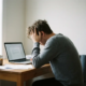 Freelancer leunt uitgeput op hand aan minimalistische thuiswerkplek met laptop, verspreide papieren en onaangeroerde koffie.