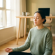 Freelancer zit gekruisbeens op houten vloer in meditatiehouding, ogen gesloten, handen op knieën, thuiskantoor op de achtergrond.