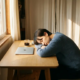 Freelance ondernemer slapend aan een minimalistische eiken bureau, hoofd op gevouwen armen naast laptop en koffiekop, warm ochtendlicht.