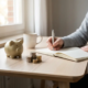 Freelancer aan modern houten bureau met spaarpot en gestapelde euromunten naast een opengeslagen notitieboek, warm natuurlijk licht.