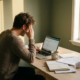 Freelancer leunt vermoeide op hand aan minimalistische houten thuiswerkplek met laptop, notitieboeken en koud kopje koffie.