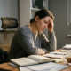 Freelancer uitgeput in ergonomische bureaustoel, hand tegen slaap, rommelig bureau met notitieboeken en vergeten koffie in zacht middaglicht.