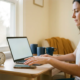 Freelancer aan minimalistisch thuiskantoor bereikt naar smartphone naast laptop, analoge klok benadrukt urgentie in warm ochtendlicht.