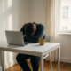 Uitgeputte freelancer met hoofd op gevouwen armen aan wit bureau, gesloten laptop en onaangeroerd kopje koffie, zacht natuurlijk licht.
