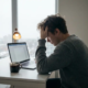 Freelancer leunt vermoeid op hand voor laptop met onaangeroerde koffie in grijs ochtendlicht.