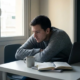 Freelancer met gespannen schouders starend naar laptop op wit bureau, omringd door notitieboeken en onaangeroerde koffie, grijs ochtendlicht.