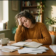 Uitgeputte freelancer leunt met hoofd in hand aan rommelig houten bureau, omringd door notitieboek en papieren in warm middaglicht.