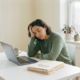 Vermoeide freelancer leunt op hand aan wit bureau met halfopen laptop, medische documenten en koffiekop in zacht daglicht.
