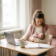 Zelfstandige moeder die haar baby voedt aan een Scandinavisch thuiskantoor bureau, laptop en koffie binnen handbereik, zacht natuurlijk daglicht.