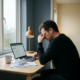 Uitgeputte ondernemer aan minimalistisch thuiskantoor, hoofd in hand, omringd door financiële documenten en laptop bij overcast raamlicht.