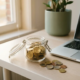 Klein glazen potje gevuld met gouden euromunten op een modern bureau naast een laptop, warm middaglicht.
