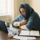 Zieke freelancer aan thuiswerkplek met laptop, dampende thee en deken om schouders, onafgewerkte papieren op bureau.