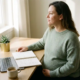 Zwangere freelancer aan een minimalistisch thuiskantoor met laptop, notitieboek vol berekeningen, plant en koffie in warm daglicht.