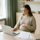 Zwangere freelancer aan Scandinavisch houten bureau met laptop en klantdocumenten, hand op buik, zacht natuurlijk daglicht.