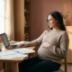 Zwangere freelancer aan houten bureau met laptop en documenten, hand op buik, warm natuurlijk licht.