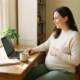 Zwangere freelancer werkt thuis aan laptop, hand op buik, met planner en thee op bureau in warm natuurlijk licht.