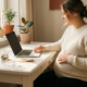 Zwangere freelancer aan bureau met laptop en spaarpot met munten, hand op buik, warm middaglicht door raam.
