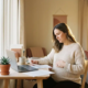 Zwangere vrouw aan thuiswerkplek bekijkt documenten met hand op buik, laptop en succulent op bureau, warm middaglicht.