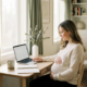 Zwangere vrouw aan modern thuiskantoor bureau met hand op buik, laptop en notitieboek, warm natuurlijk licht.