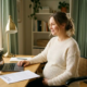 Zwangere vrouw aan thuiswerkplek met laptop, freelancematerialen en arbeidsovereenkomst in warm natuurlijk licht.